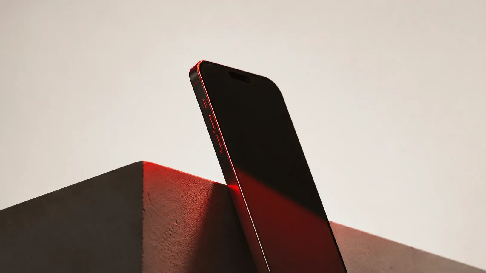 Ein schwarzes Smartphone lehnt an einem dunklen Betonblock; warmes orangefarbenes Licht streift seitlich die Gerätekante und den Beton, der Hintergrund bleibt schlicht hellgrau.